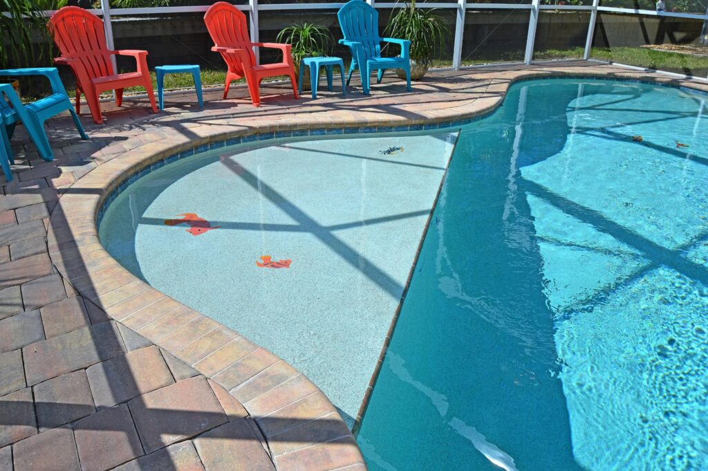 A shallow beach-entry pool shelf with fish decals, surrounded by colorful chairs under a screened enclosure.