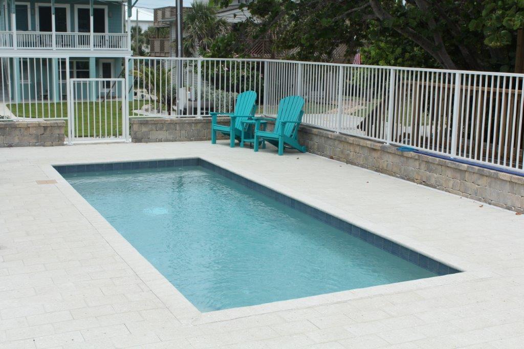 A small rectangular pool with dark tile edging on a paver patio, enclosed by a white metal fence.