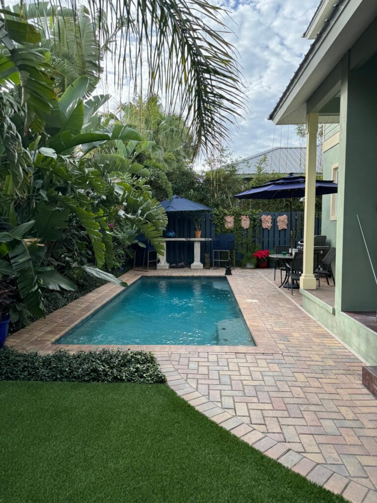 A small rectangular backyard pool lined with brick pavers and lush tropical landscaping beside a house.