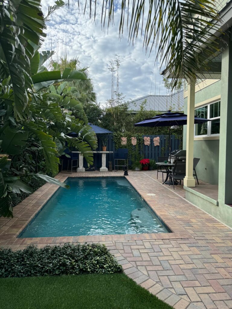 A small rectangular backyard pool with brick paver decking surrounded by tropical palm trees and a patio dining area.