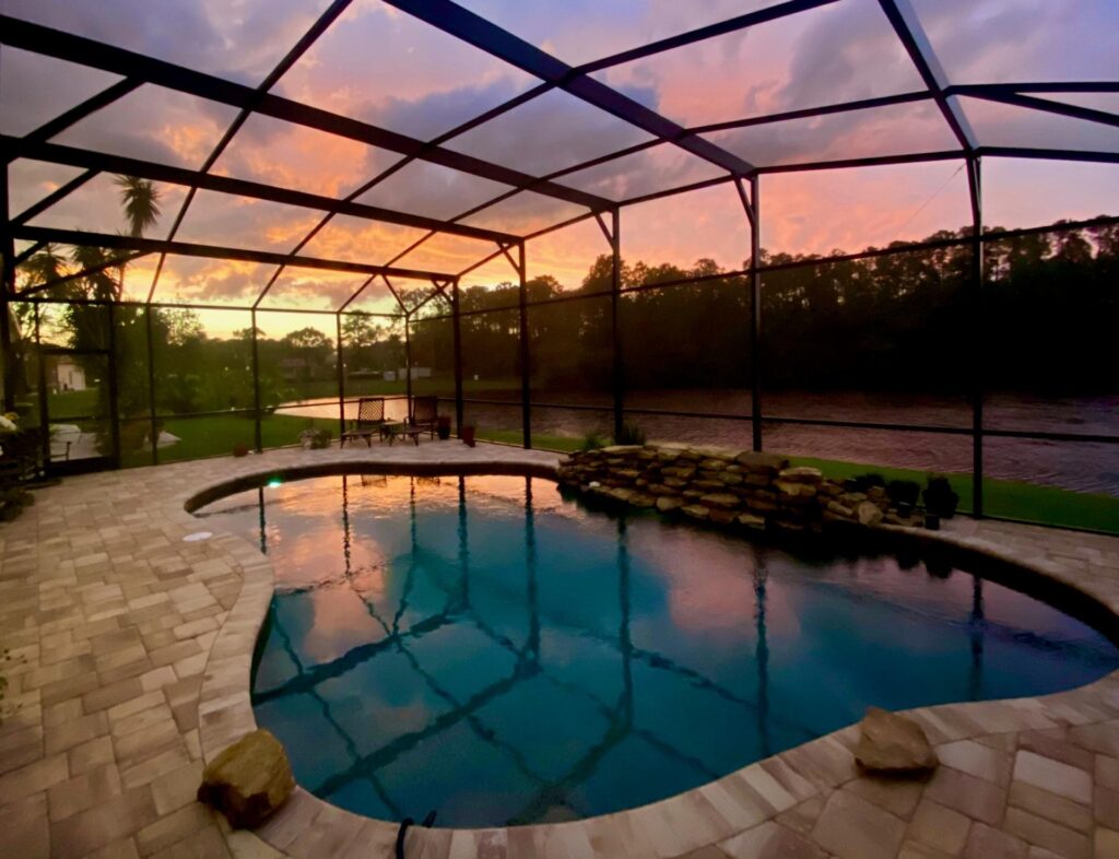 A freeform pool with a rock waterfall under a screened enclosure at sunset.
