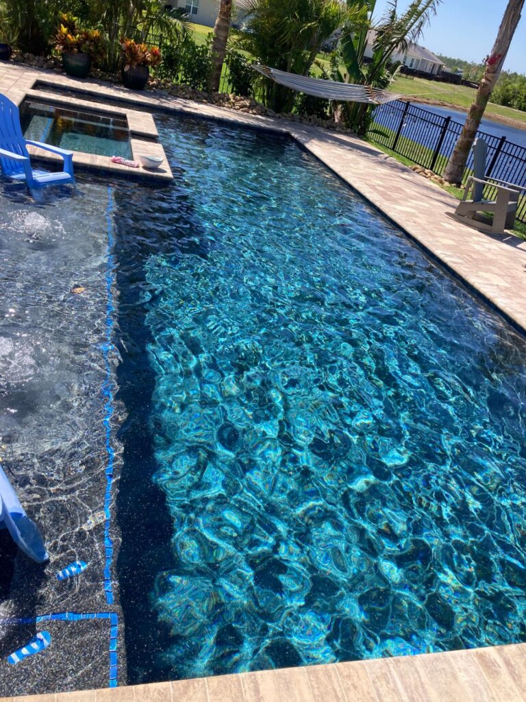 A rectangular pool with a raised spa and blue pebble-finished waterline, overlooking a canal with a paver deck and palm trees.