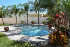 A freeform backyard pool surrounded by palm trees and a paver patio with a white vinyl fence.
