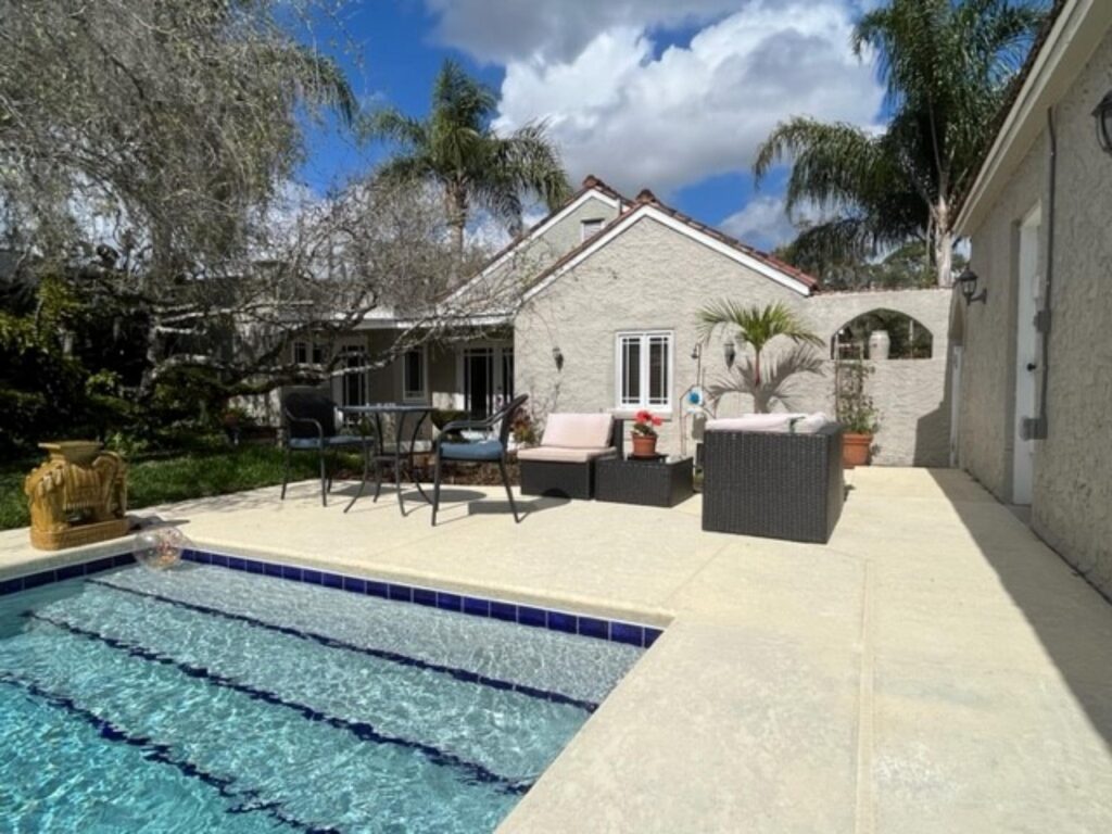 A corner of a backyard pool with blue tile trim, next to a patio seating area and Spanish-style house.