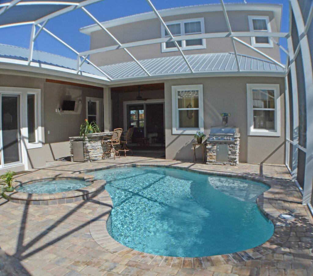 A freeform pool with an attached raised spa sits under a screened lanai next to an outdoor kitchen.