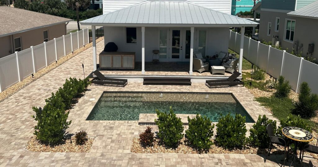 Small rectangular backyard pool with paver deck, lounge chairs, and covered porch behind it.