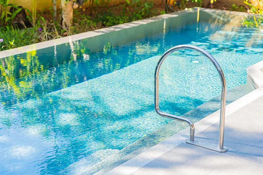 A stainless steel handrail beside a narrow blue lap pool with tiled edge and greenery beyond.