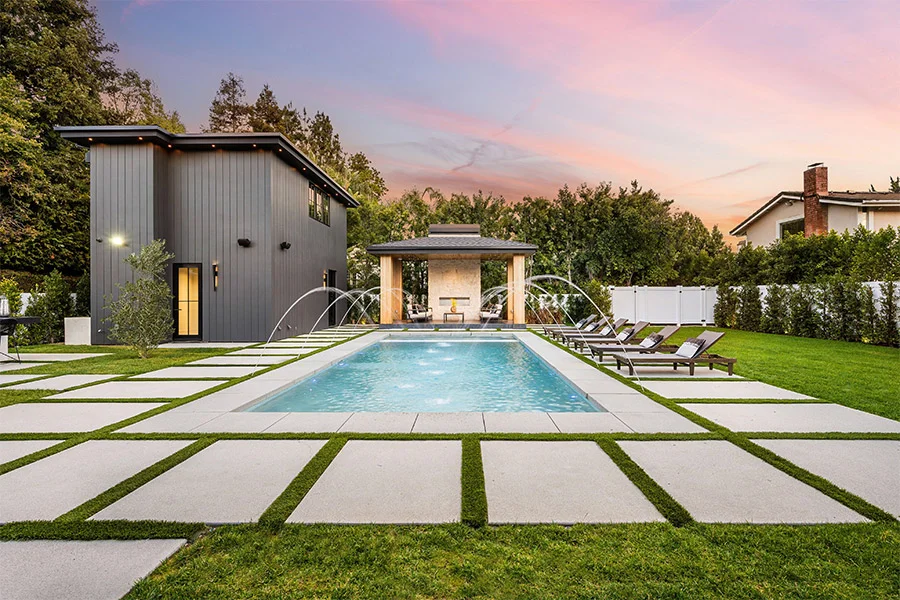 A rectangular backyard pool with arcing water jets at dusk, framed by a modern cabana and paver walkways.