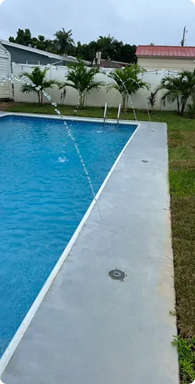 A small rectangular vinyl pool with deck jets spraying water, surrounded by a concrete patio and privacy fence.