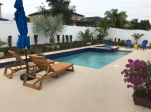 A rectangular backyard pool with a dark tiled spa in the corner, surrounded by a concrete patio and white privacy fence.