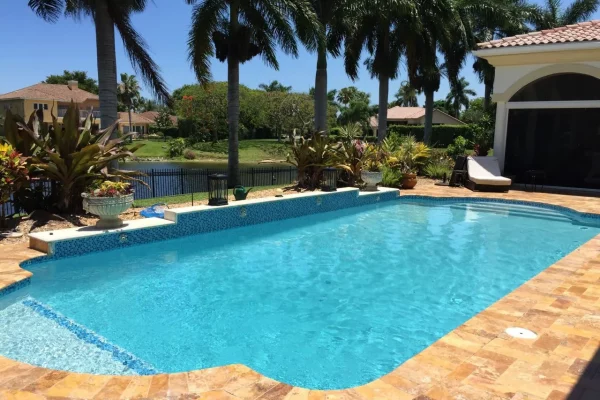 A rectangular backyard pool with travertine decking overlooking a canal lined with palm trees.