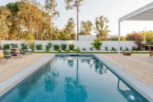 A rectangular backyard pool with light coping, wood-look decking, loungers, and a low white wall lined with shrubs.