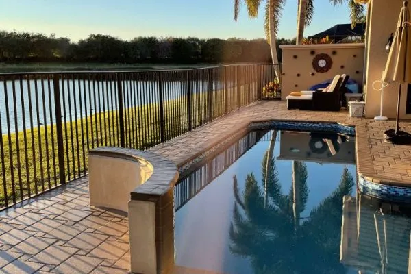 A pool corner at dusk with a paver deck overlooking a lake behind a metal fence.