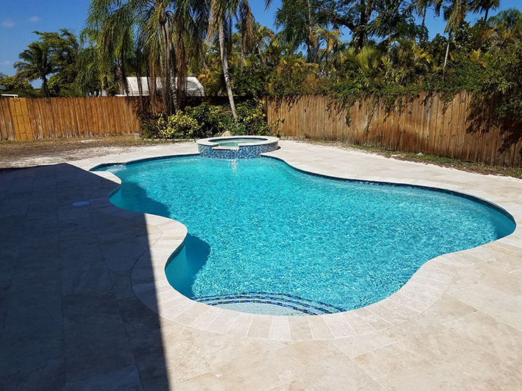 A freeform pool with a raised spa surrounded by travertine pavers and a wooden privacy fence.