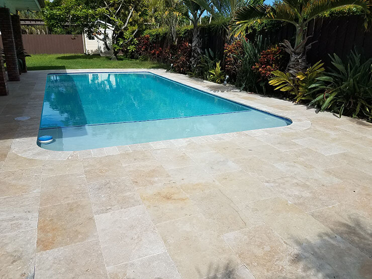 A rectangular backyard pool with turquoise water and a large travertine paver patio surrounded by tropical landscaping.