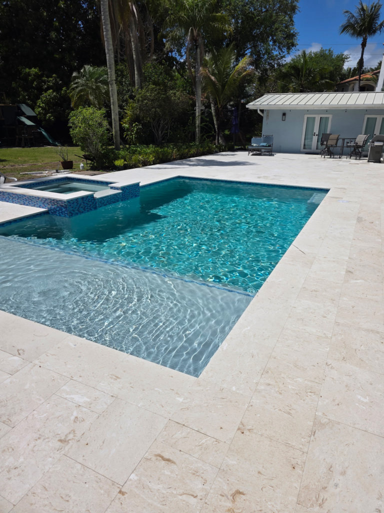 A rectangular pool with a sun shelf and raised spa on a travertine patio behind a house.