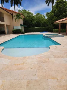 A freeform rectangular pool with a shallow rounded end and an attached raised spa, set on a travertine paver patio behind a large house.