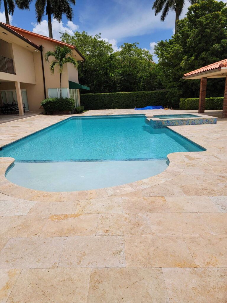 A freeform rectangular pool with a shallow rounded end and an attached raised spa, set on a travertine paver patio behind a large house.