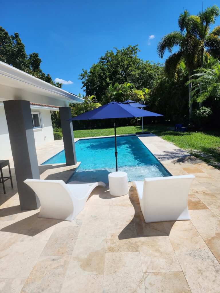 A small rectangular backyard pool with turquoise water beside a travertine patio with white lounge chairs and a blue umbrella.