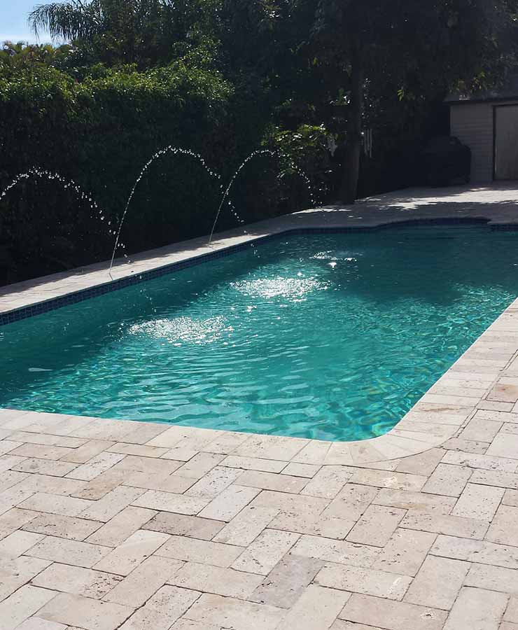 A small rectangular backyard pool with three arcing deck jets and travertine paver decking.