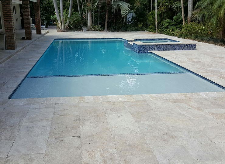 A rectangular gunite pool with a raised spa and blue tile waterline surrounded by travertine pavers and tropical landscaping.