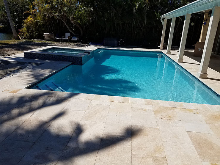 A rectangular backyard pool with a raised spa and travertine paver deck under a covered patio.