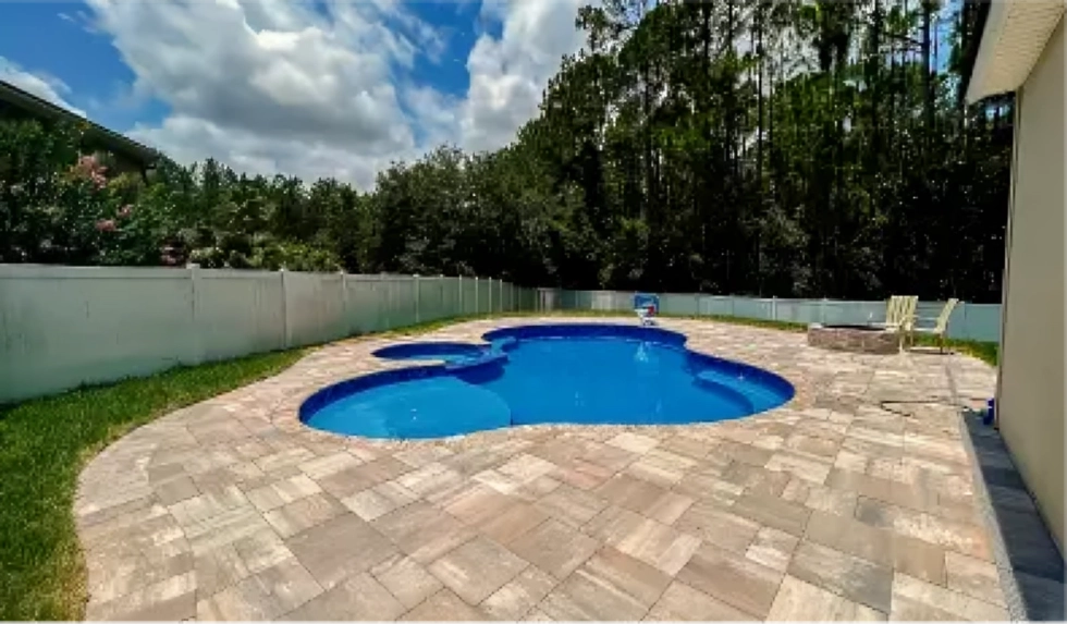 A freeform fiberglass pool with an attached round spa surrounded by paver decking and a white privacy fence.