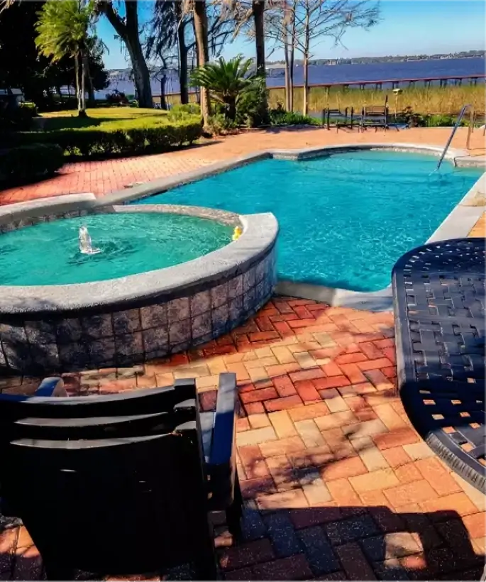 A freeform pool with a raised stone spa and fountain jet sits on a brick paver patio overlooking a lake.