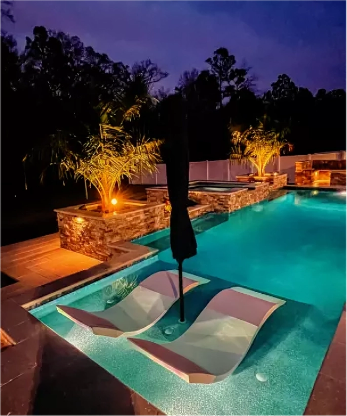 Two in-pool loungers with an umbrella sit on a sun shelf at night, lit turquoise, with a raised stone spa behind.