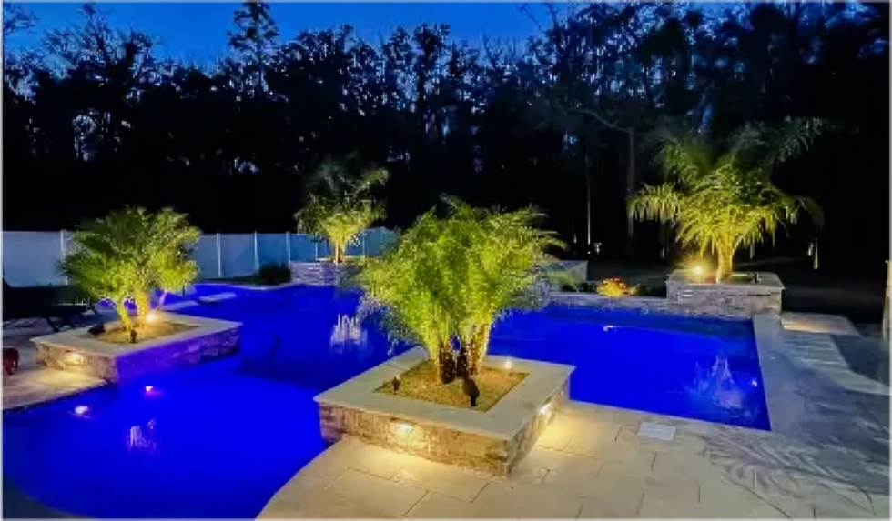 A freeform backyard pool lit blue at night with raised stone planters holding palm trees.