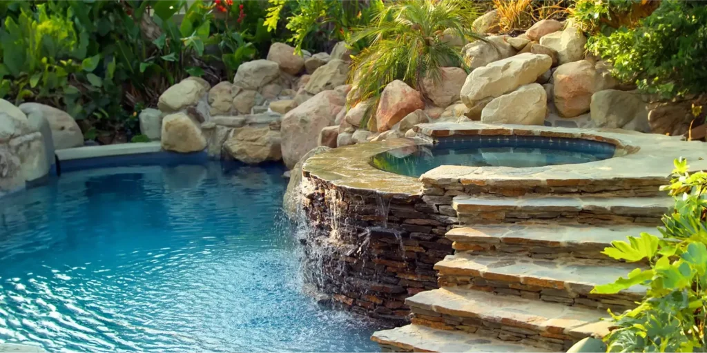 A raised stone spa spills a waterfall over stacked rock into a freeform pool surrounded by boulders and lush plants.