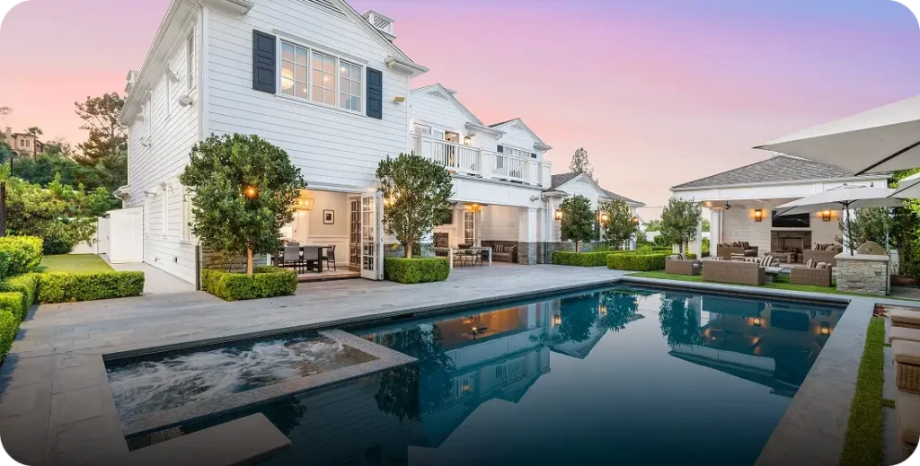 A rectangular backyard pool with a raised spillover spa reflects a pastel dusk sky beside a large white house.