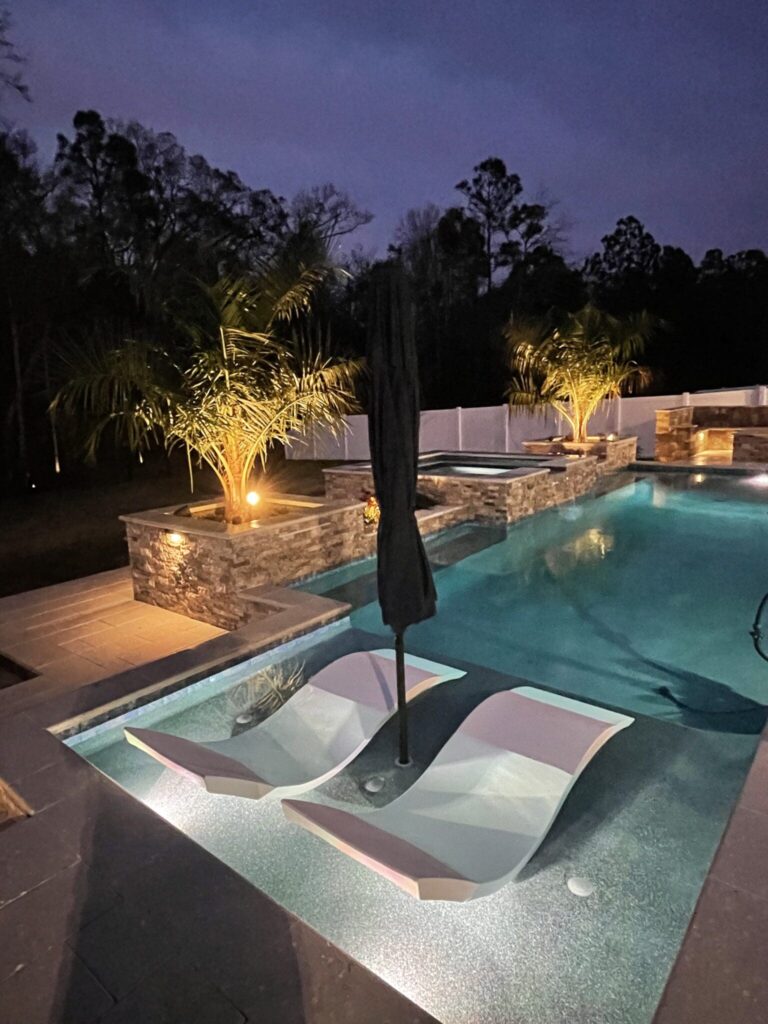 Rectangular pool at night with two built-in loungers and a raised stone spa in the background.