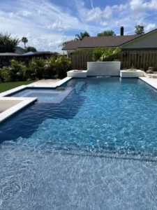 A rectangular pool with a raised spa, tanning ledge, and a sheer descent water wall feature under a bright blue sky.