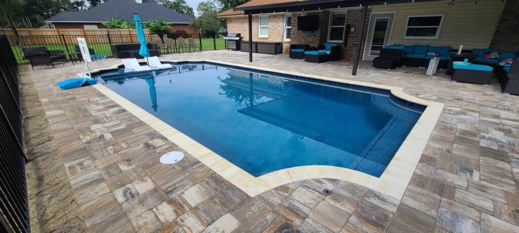 A rectangular vinyl pool with tan coping on a paver patio behind a house with covered lanai seating.