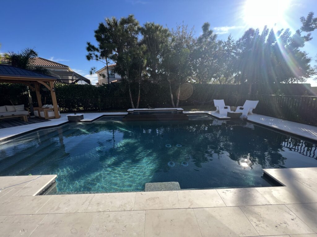 A dark-bottomed rectangular pool with a raised spa area and travertine deck under a sunny sky.