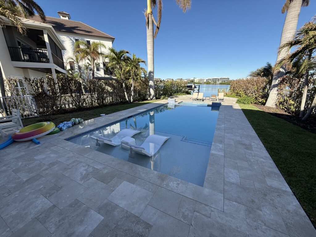 A rectangular pool with in-water loungers and a raised spa overlooks a canal lined with palm trees.