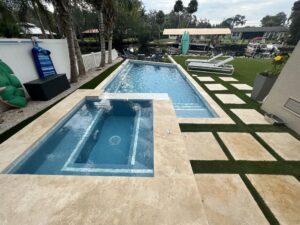 A rectangular pool with an attached raised spa on a paver deck beside a canal lined with palm trees.