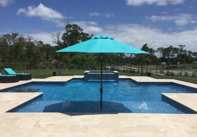 A rectangular pool with a raised spa spillover and tanning ledge under a large umbrella on a travertine patio.