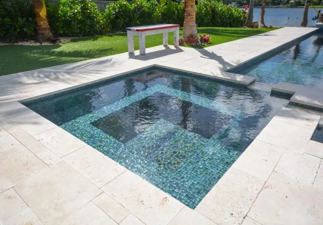 A raised square spa with vivid blue-green tile sits beside a narrow lap pool on a travertine patio near the water.