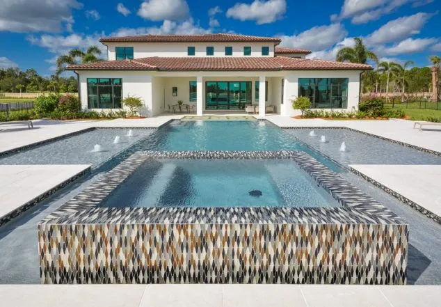 A modern geometric pool with a raised spa faced in colorful mosaic tile in front of a large white stucco house.