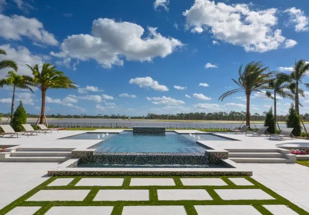 A modern multi-level rectangular pool with a raised water-jet wall and lower reflecting basin, set on a paver-and-turf patio overlooking open water.