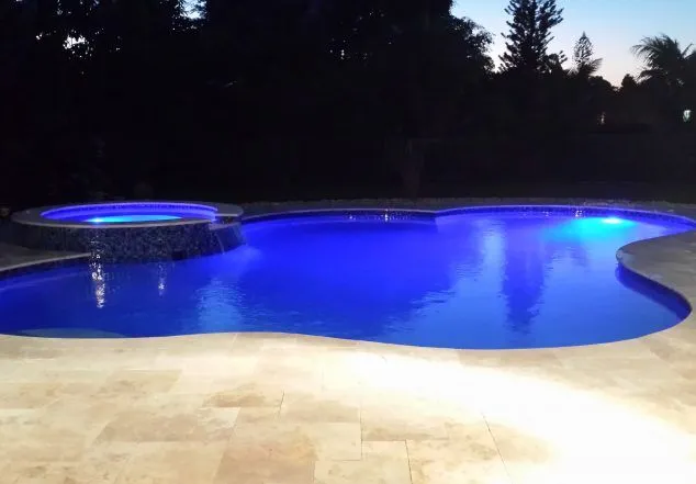 A freeform pool with a raised spillover spa lit by blue LED lighting at night.