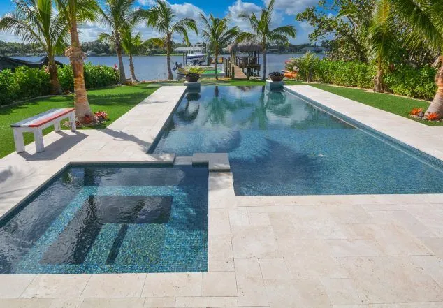 A large rectangular infinity-edge pool with a raised spa overlooks a canal lined with palm trees and a private dock.