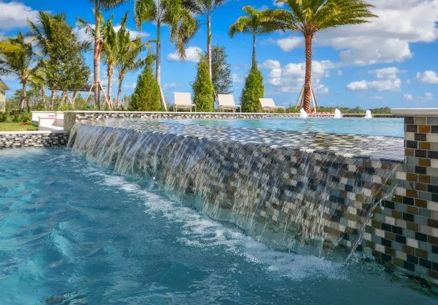 A tiled raised wall cascades water into a pool with palm trees and a lake in the background.
