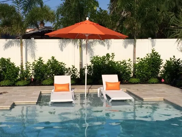 Two white loungers with orange cushions sit on a sun shelf in a pool under an orange umbrella.