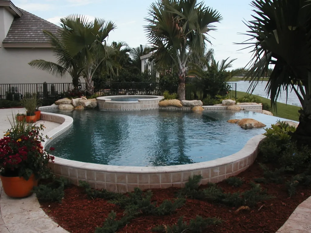 A freeform pool with a raised rock-accented spa sits on a canal-front backyard lined with palm trees.