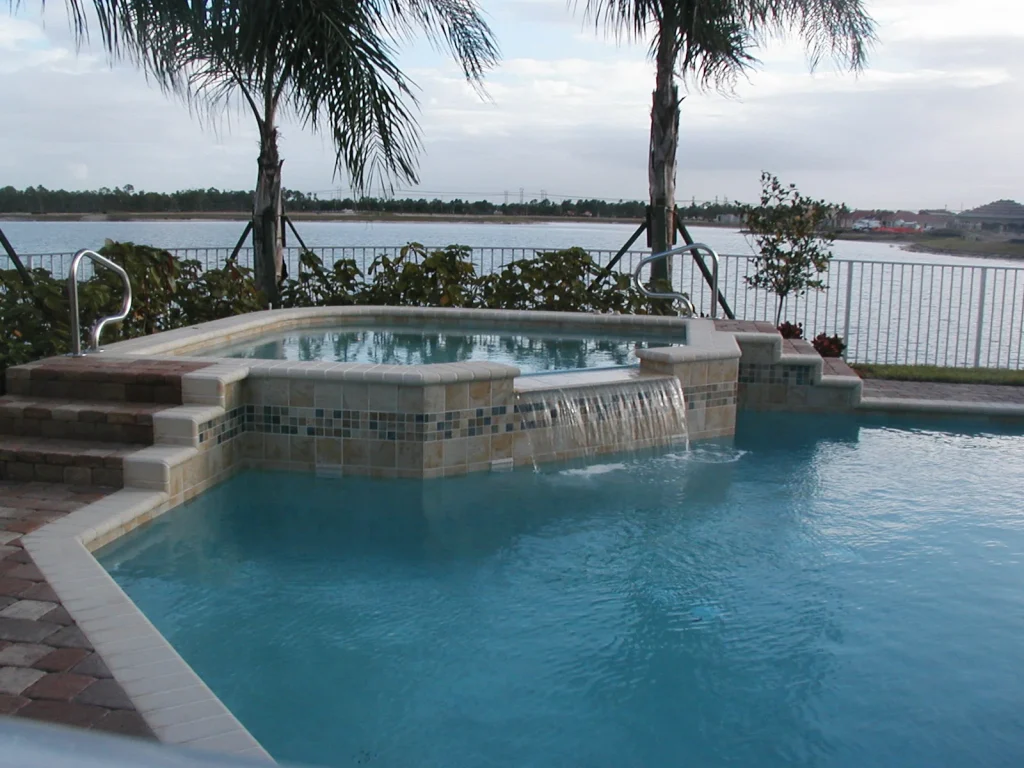 A raised spa with tiled wall spills water into a pool overlooking a lake.