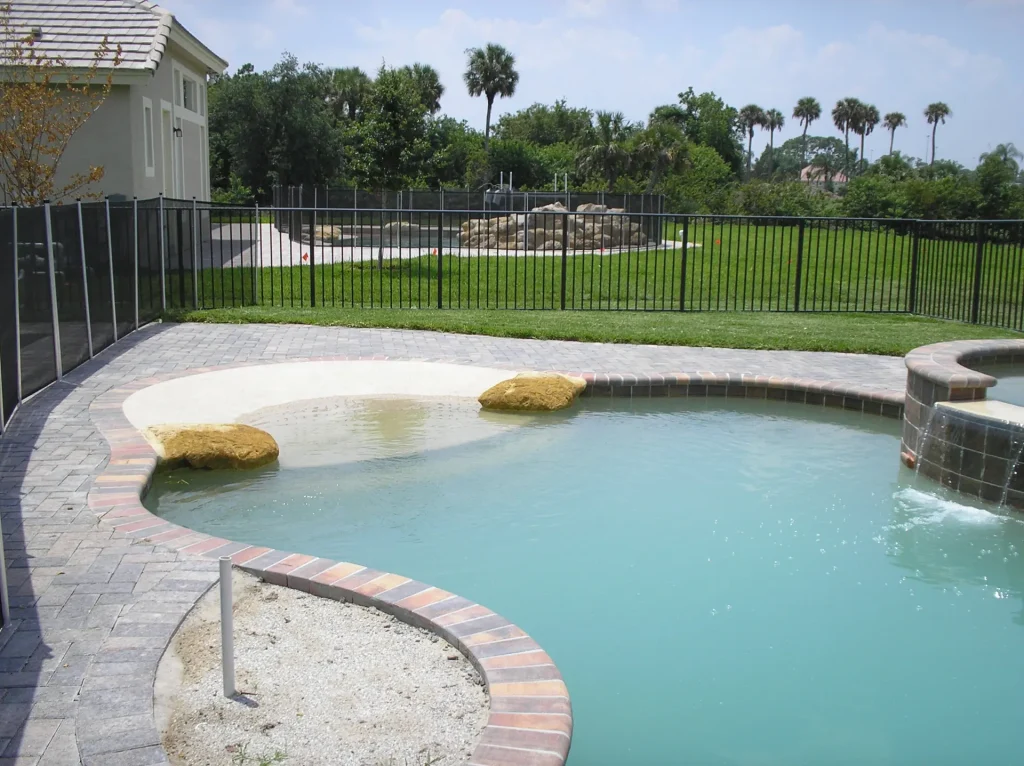 A freeform pool with a raised spa spillover and beach entry, enclosed by a black metal fence with a grassy backyard beyond.