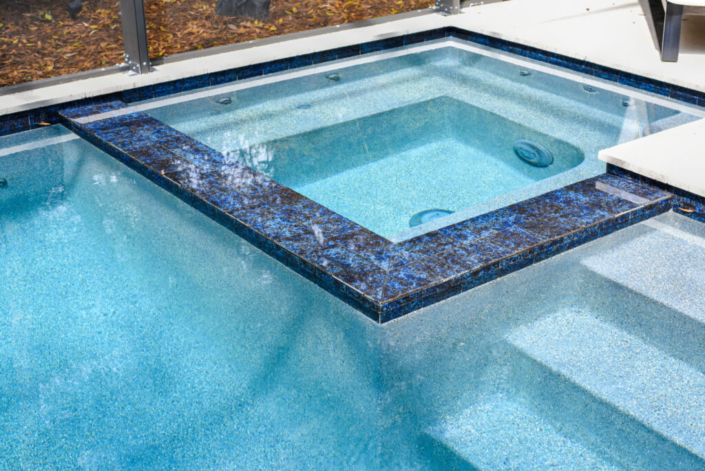 Close-up of a raised spa with dark blue mosaic tile edge overlooking a speckled-finish pool with entry steps.